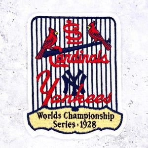 May include: Embroidered patch featuring the Cardinals and Yankees logos, with two red birds perched on a branch. The patch has a striped background and text that reads "Worlds Championship Series 1928".