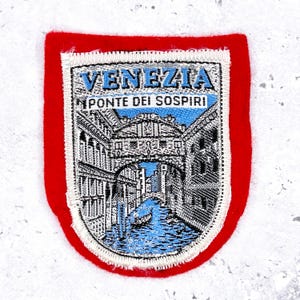 May include: Embroidered patch depicting the Bridge of Sighs in Venice, Italy, with the city and bridge names in blue. The patch has a white border and a red felt backing, a classic travel souvenir. The design is detailed.