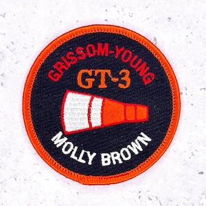 Peut inclure: Un écusson rond brodé avec le texte "GRISSOM-YOUNG GT-3" en rouge et "MOLLY BROWN" en blanc. L'écusson présente un fond noir, une bordure orange et un graphisme stylisé orange et blanc.