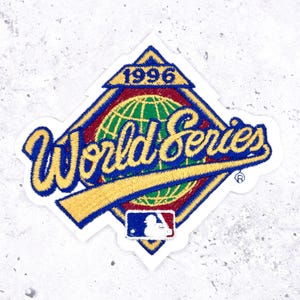 May include: Embroidered patch featuring the words "World Series" in a yellow script font, with a blue outline. The patch has a diamond shape with a globe in the center, the year "1996" at the top, and the MLB logo at the bottom.