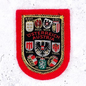 May include: A rectangular red felt patch featuring a black embroidered centre with gold trim. The design displays the text OSTERREICH AUSTRIA and various Austrian state coats of arms, including labels like N.OST, BGLD, WIEN, VLBG, TIROL, KTN, SBG, ST, and MK.