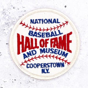 Peut inclure: Un écusson brodé circulaire sur fond blanc cassé avec des motifs de coutures de baseball rouges. Le texte indique NATIONAL BASEBALL HALL OF FAME AND MUSEUM COOPERSTOWN N.Y. en fil bleu et rouge avec une bordure cousue finie.