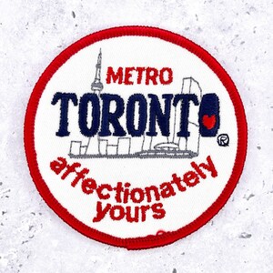 May include: Embroidered patch with a white background and a red border. The patch features the word "TORONTO" in navy blue, the word "METRO" in red, and the phrase "affectionately yours" in red. A heart is included.