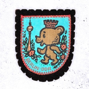 Könnte beinhalten: Ein Vintage-Patch mit einem Cartoon-Bären mit Krone, dem Berliner Fernsehturm und dem Text "BERLIN DDR". Der Patch hat einen schwarzen, gewellten Rand, einen rot genähten Rand und einen türkisfarbenen Hintergrund.