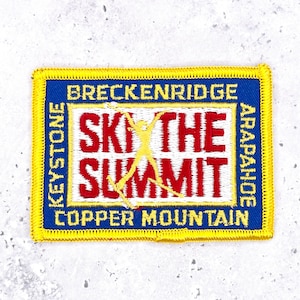 May include: A rectangular embroidered patch with a yellow border and blue background. The center features the red text 'SKI THE SUMMIT' with a yellow skier silhouette. The border text reads 'BRECKENRIDGE', 'KEYSTONE', 'ARAPAHOE', and 'COPPER MOUNTAIN'.