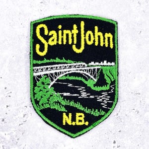 Könnte beinhalten: Gestickter Aufnäher mit den Worten "Saint John" in gelber Schrift über einer Brückenillustration. Der Aufnäher hat einen schwarzen Hintergrund mit grünen Akzenten und den Buchstaben "N.B." unten.