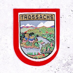 Pode incluir: Remendo bordado com uma borda vermelha e uma cena de paisagem detalhada. O remendo apresenta a palavra "TROSSACHS" no topo. A paisagem inclui um rio, árvores e montanhas, com um fundo de céu azul.