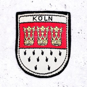 以下が含まれることがあります： 「KÖLN」の文字が刺繍されたパッチ。赤色のストライプの上に3つの金の王冠が描かれています。その下には、黒い涙型のシンボルが配置された白いフィールドがあります。パッチは黒い縁取りがされています。