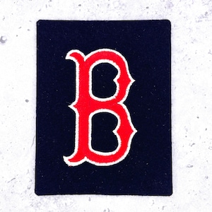 May include: A rectangular black fabric patch featuring a large, stylized letter B embroidered in bright red thread with a white border. The embroidery has a dense, textured finish, and the letter is centered on the dark, solid-colored background.