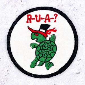 May include: Embroidered patch featuring a green turtle wearing a black top hat and red scarf. The turtle is walking and has a patterned shell. The text "R-U-A-?" is above the turtle in red letters. The patch has a white background and a black border.