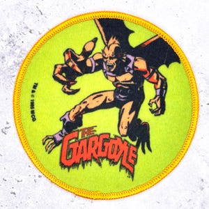 Könnte beinhalten: Runder Aufnäher mit einer Cartoon-Figur einer Gargoyle in Orange-, Lila- und Schwarztönen auf einem limettengrünen Hintergrund. Der Text "THE GARGOYLE" ist orangefarben, mit einem gelben Rand. Gelber, genähter Rand.