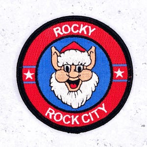 May include: Embroidered patch with a cartoon elf design. The patch features a red and blue color scheme with the words "ROCKY ROCK CITY" in white. Two white stars are on the red stripes.