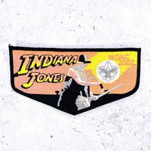 May include: Embroidered patch featuring the words "INDIANA JONES" in yellow and black, with an image of the character. The patch also includes "NOAC 2002" and a Boy Scouts of America emblem. The patch has a black border and a peach and yellow background.