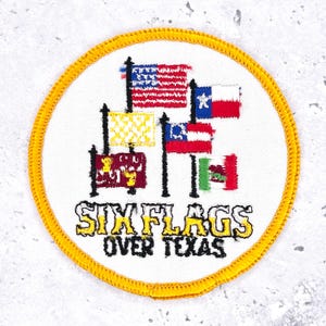 May include: Embroidered patch featuring the text "SIX FLAGS OVER TEXAS" in yellow and black. The patch displays several flags, including the American flag and the Texas flag, on a white background with a yellow border.