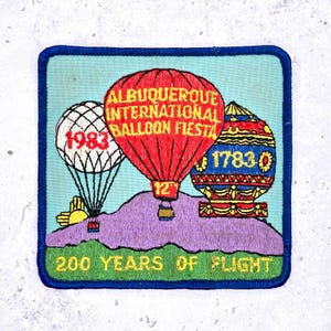 May include: Embroidered patch featuring hot air balloons and the text "Albuquerque International Balloon Fiesta." The patch includes the years 1983 and 1783, with the phrase "200 Years of Flight." The patch has a blue border and a light blue background.