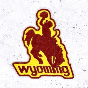 May include: Embroidered patch featuring a maroon bucking bronco and rider on a yellow background. The word "WYOMING" is embroidered in yellow at the bottom. The patch has a unique shape, with a curved top and a rectangular base.