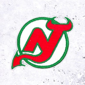 May include: Embroidered patch featuring the logo of the New Jersey Devils hockey team. The logo is a red "N" with devil horns and tail, set within a white circle, and outlined with green stitching.