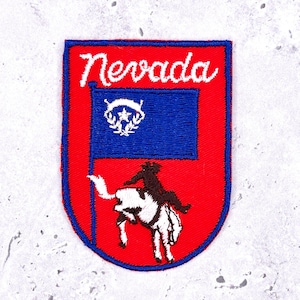 May include: Embroidered patch featuring the word "Nevada" in white script above a blue flag with a white emblem. Below the flag is a black silhouette of a rider on a bucking horse, set against a red background.