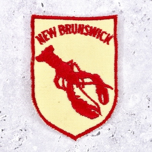 May include: A rectangular patch with a cream-colored background and a red embroidered lobster design. The words "NEW BRUNSWICK" are arched above the lobster. The patch has a red border.