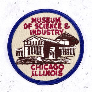Op de afbeelding: Een ronde geborduurde patch met een crèmekleurige achtergrond en een donkerblauwe rand. Het toont een donkerrode geborduurde illustratie van het Museum of Science & Industry-gebouw met de tekst MUSEUM OF SCIENCE & INDUSTRY CHICAGO ILLINOIS.