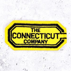 May include: A rectangular embroidered patch with clipped corners, featuring a bright yellow background and a thick black border. The text THE CONNECTICUT COMPANY is stitched in black capital letters across the center of the patch.