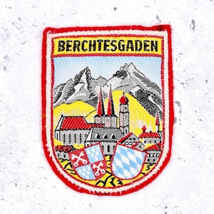 May include: Embroidered patch with a red border and the word "BERCHTESGADEN" in yellow. The patch features a mountain range, buildings, and two shields with heraldic designs. The patch is a souvenir or emblem.