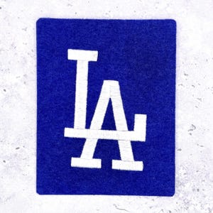 Puede incluir: Un parche de fieltro azul con las letras blancas bordadas "LA". El parche es rectangular con esquinas redondeadas. Las letras están en una fuente estilizada, comúnmente asociada con un equipo deportivo.