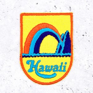 May include: Embroidered patch with a yellow background and an orange border. The patch features a rainbow design in orange, blue, and turquoise, with the word "Hawaii" in turquoise script. The patch is a shield shape.