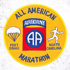 May include: A circular yellow patch with the words "ALL AMERICAN AIRBORNE MARATHON" in blue. It features a red and blue emblem with "AA" and a white parachute and runner. Text includes "FORT BRAGG" and "NORTH CAROLINA."