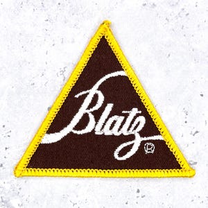 May include: A triangular embroidered patch with a dark brown background and a bright yellow stitched border. The word Blatz is written in white cursive script across the centre, with a small registered trademark symbol in the bottom right corner.