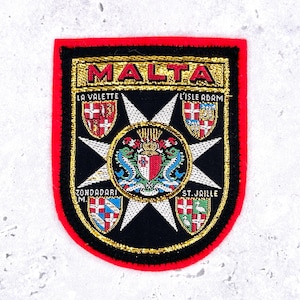 May include: A rectangular embroidered patch with a red border and black background featuring the word MALTA in gold lettering. It displays a central white eight-pointed cross, a circular crest, and four smaller heraldic shields labeled with text.