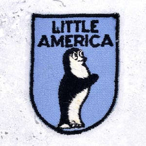 May include: A shield-shaped embroidered patch with a light blue background and a black stitched border. The text LITTLE AMERICA is embroidered in black at the top. Below the text is a black and white embroidered figure of a standing penguin.