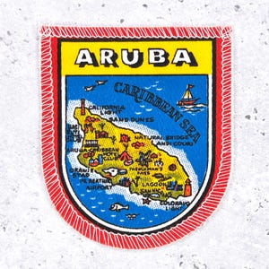 May include: A rectangular Aruba patch with a red border and white stitching. The patch features a yellow top section with the word "ARUBA" and a blue lower section with a map of the island and the words "CARIBBEAN SEA".