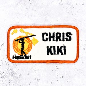 May include: A rectangular embroidered patch with an orange border and white background. It features a black silhouette of a person carrying a pole over a yellow sun, a yellow map of Hawaii, and the black text CHRIS KIKI and Hawaii.