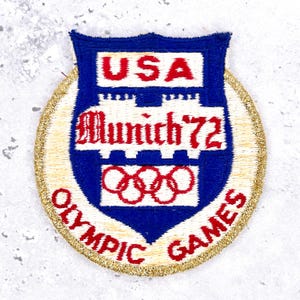 May include: A circular embroidered patch featuring a blue shield shape on a cream background. The text reads USA, Munich '72, and Olympic Games in red. Five red Olympic rings are centered. The border is finished with metallic gold thread.
