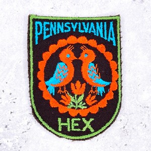 Op de afbeelding: Een schildvormige geborduurde patch met een zwarte achtergrond en een felgroene rand. Het bevat de tekst PENNSYLVANIA in blauw aan de bovenkant en HEX in groen aan de onderkant. Twee oranje vogels met blauwe vleugels staan tegenover elkaar in een oranje cirkel.