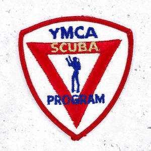 Può includere: Toppa bianca e rossa con la scritta "YMCA SCUBA PROGRAM". La toppa presenta un triangolo rosso con una silhouette di subacqueo blu. La toppa ha un bordo rosso.
