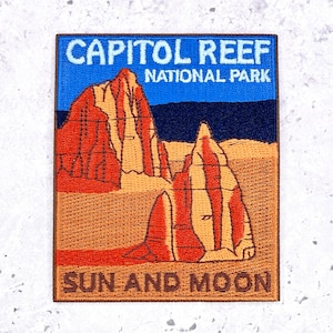 May include: A rectangular embroidered patch featuring a landscape of Capitol Reef National Park with red and tan rock formations against a blue sky. The text reads CAPITOL REEF NATIONAL PARK at the top and SUN AND MOON at the bottom in brown.