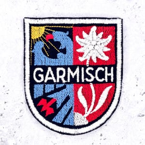 May include: A shield-shaped embroidered patch featuring a split design. The left side is light blue with a yellow sun and a red bird. The right side is red with a white edelweiss flower and white plant stems. The word GARMISCH is in white text.