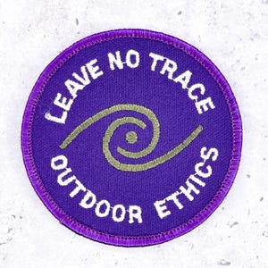 May include: A circular purple embroidered patch featuring a central olive green spiral design. The text "LEAVE NO TRACE" is arched across the top and "OUTDOOR ETHICS" is arched across the bottom in white, bold, capitalized, sans-serif lettering.