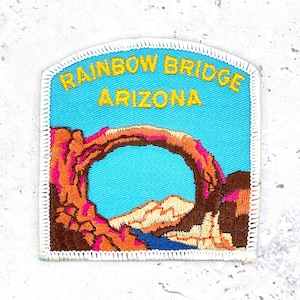 May include: Embroidered patch featuring the Rainbow Bridge in Arizona. The patch has a blue background with the words "RAINBOW BRIDGE ARIZONA" in yellow. The bridge is depicted in shades of brown, orange, and pink.