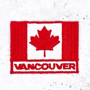 May include: Embroidered patch depicting the Canadian flag. The design includes a red maple leaf on a white square, with red bars on either side. The word "VANCOUVER" is embroidered in red below the flag.