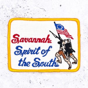 May include: A rectangular embroidered patch with a yellow border on a white background. It features the red text Savannah and blue text Spirit of the South. To the right is an embroidered figure holding a rifle and a red, white, and blue flag.