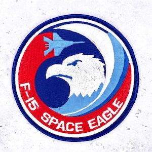May include: Embroidered patch with a circular design featuring a white eagle head, a blue jet, and the text "F-15 SPACE EAGLE." The patch has a red and blue color scheme with white accents and a blue border.