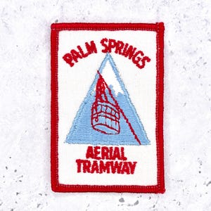 May include: A rectangular embroidered patch with a white background and a red border. It features a light blue triangle containing a red tram car graphic. The text reads PALM SPRINGS in red at the top and AERIAL TRAMWAY in red at the bottom.