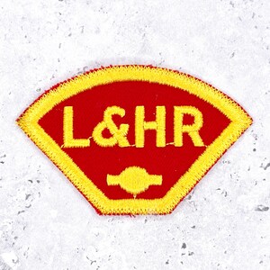 May include: A fan-shaped embroidered patch featuring a vibrant red background with a bright yellow border. The letters L&HR are stitched in yellow across the center, positioned above a small, solid yellow circular icon with two horizontal extensions.