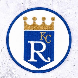May include: A circular embroidered patch featuring a white background with a blue border. Centered is a blue shield topped with a gold crown. Inside the shield is a large white letter R and the smaller gold letters KC positioned above it.