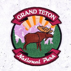 May include: A circular embroidered patch featuring a brown moose standing in a green forest. Behind the moose are light purple mountains and an orange sunburst. The text GRAND TETON is on a red banner above, and National Park is on a red banner below.
