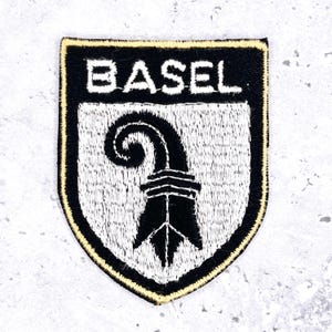 May include: Embroidered patch featuring the word "BASEL" in white capital letters above a black and white emblem. The emblem is a stylized staff with a curved top. The patch has a black border with a gold trim.