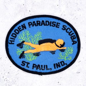 May include: An oval embroidered patch with a bright blue background and a black border. It features a yellow scuba diver, green seaweed, and black text that reads HIDDEN PARADISE SCUBA and ST. PAUL, IND. The design is centered on the blue fabric.